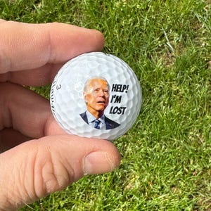 Puede incluir: Una bola de golf blanca con una imagen de un hombre en ella. El hombre lleva un traje azul y una corbata. El texto "HELP! I'M LOST" est&aacute; impreso en la bola.
