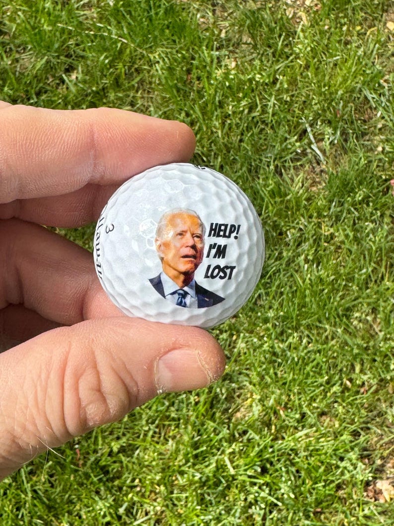 Puede incluir: Una bola de golf blanca con una imagen de un hombre en ella. El hombre lleva un traje azul y una corbata. El texto "HELP! I'M LOST" est&aacute; impreso en la bola.