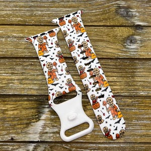 May include: White Halloween-themed watch bands featuring a repeating pattern of a character in an orange jumpsuit, pumpkins, bats, and stars. The watch bands have a white clasp and are displayed on a wooden surface.