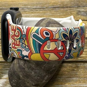 May include: A white Apple Watch band with a colorful psychedelic pattern featuring peace signs, rainbows, and flowers. The band has the word "Peace" printed on it.