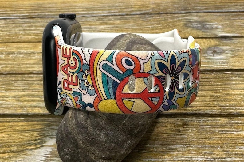 May include: A white Apple Watch band with a colorful psychedelic pattern featuring peace signs, rainbows, and flowers. The band has the word "Peace" printed on it.