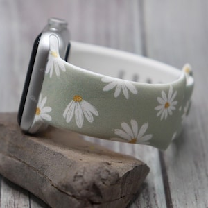 Puede incluir: Una banda de reloj Apple verde claro con estampado de margaritas blancas. La banda est&aacute; hecha de silicona y tiene un forro blanco.