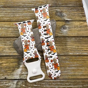 May include: Two white Halloween-themed watch bands with a repeating pattern of a character in an orange costume, pumpkins, and bats. One band is attached to a white watch connector.
