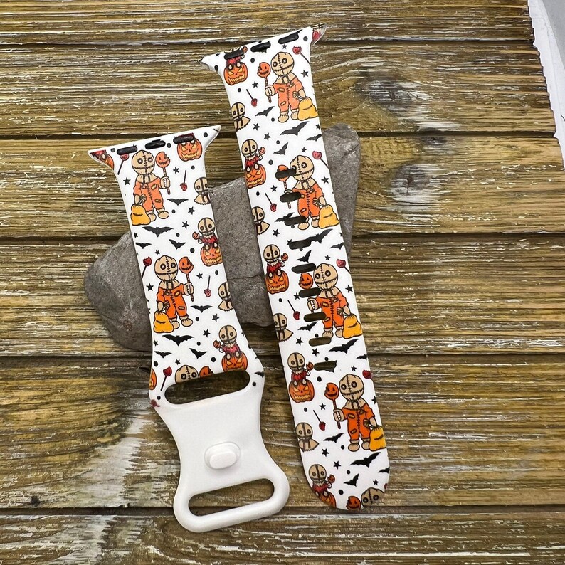 May include: Two white Halloween-themed watch bands with a repeating pattern of a character in an orange costume, pumpkins, and bats. One band is attached to a white watch connector.