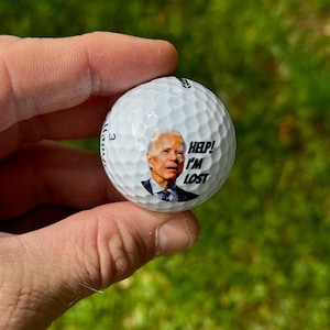 Puede incluir: Una bola de golf blanca con una imagen de Joe Biden. El texto "HELP! I'M LOST" est&aacute; impreso en la bola.
