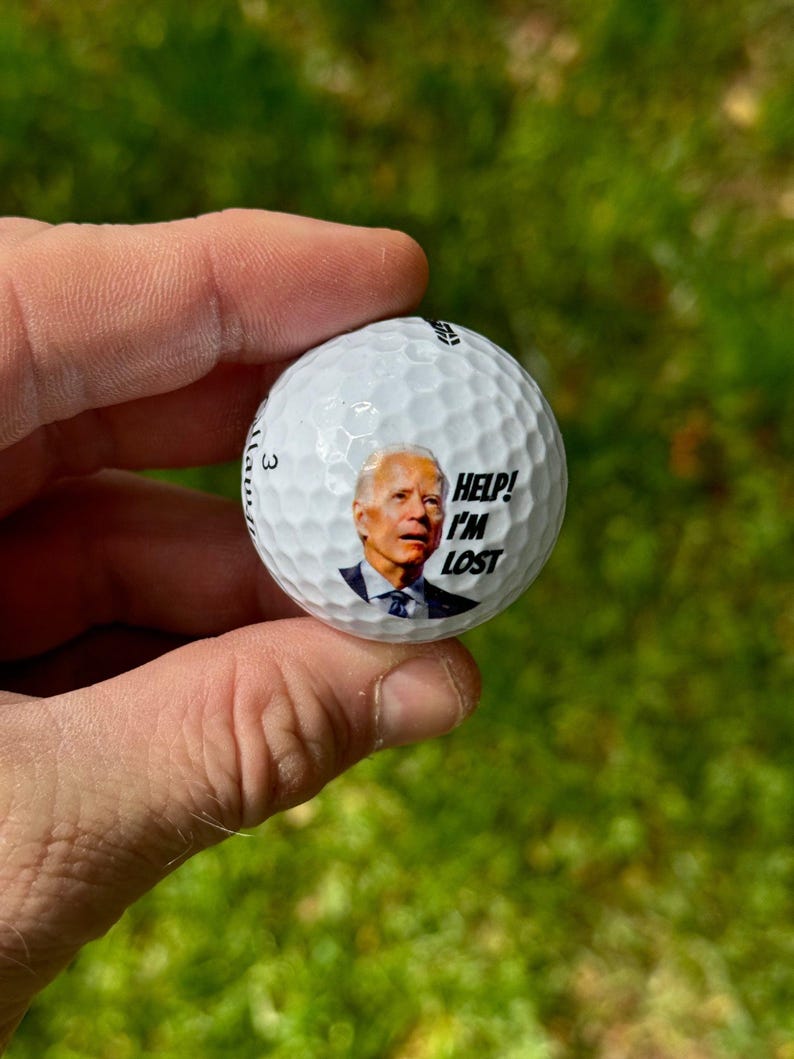 Puede incluir: Una bola de golf blanca con una imagen de Joe Biden. El texto "HELP! I'M LOST" est&aacute; impreso en la bola.