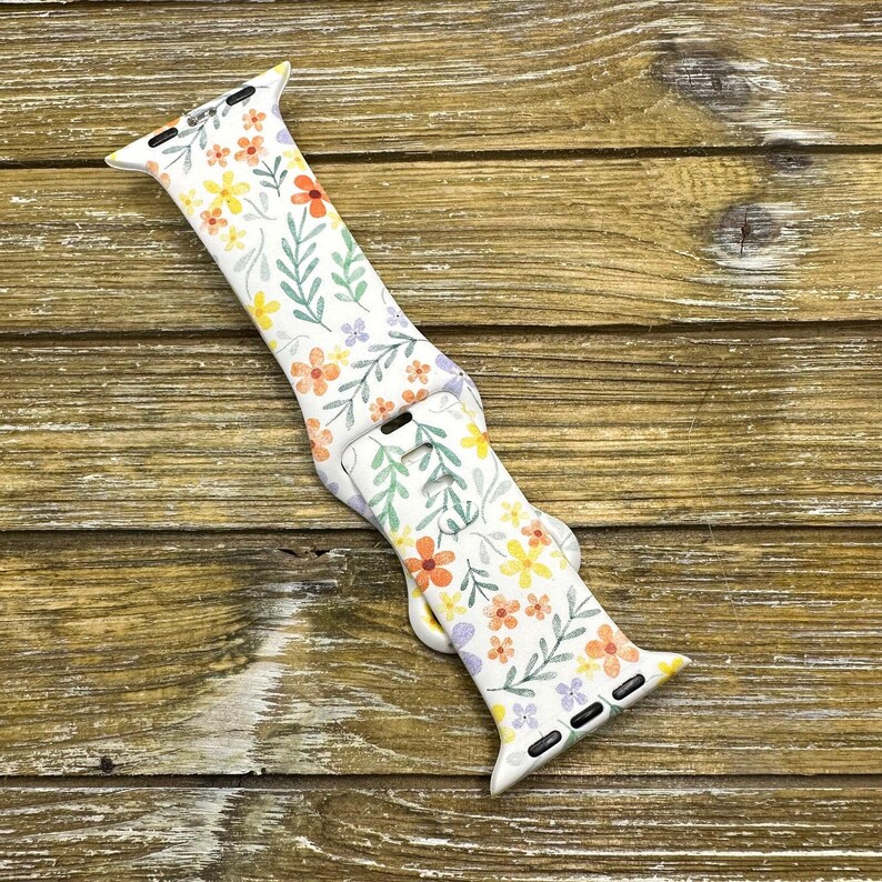 Op de afbeelding: Witte Apple Watch band met een bloemenpatroon met gele, oranje en paarse bloemen op een witte achtergrond.