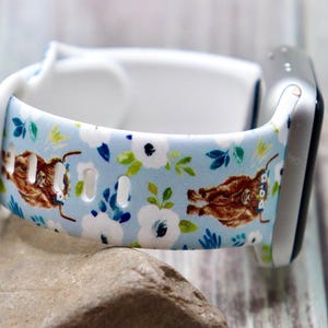 Op de afbeelding: Lichtblauwe horlogeband met een bloemen- en Schotse hooglanderpatroon. De band heeft witte bloemen, groene bladeren en bruine koeien. De horlogeband is gemaakt van een flexibel materiaal en is bevestigd aan een zilveren horloge.