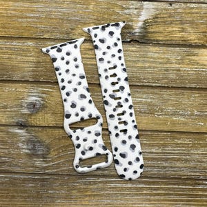 May include: Two white watch bands with a black and gray spotted pattern. The bands are made of a flexible material and have a buckle closure. The bands are designed to fit a smartwatch.
