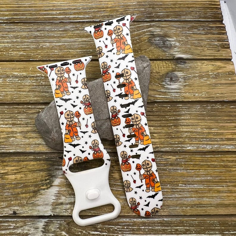 May include: Two white Halloween-themed watch bands with a repeating pattern of a character in an orange jumpsuit, pumpkins, bats, and stars. The bands are on a wooden surface.