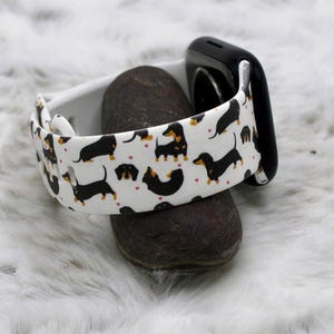 May include: A white Apple Watch band with a black and brown dachshund print. The band is on a brown rock.