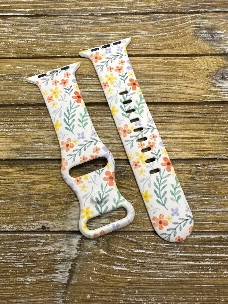 Op de afbeelding: Een wit Apple Watch bandje met een bloemenpatroon met oranje, gele en paarse bloemen op een witte achtergrond. Het bandje is gemaakt van siliconen en heeft een klassieke gespsluiting.