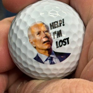 Puede incluir: Una bola de golf blanca con una imagen impresa de una persona con el texto "HELP! I'M LOST" en ella.