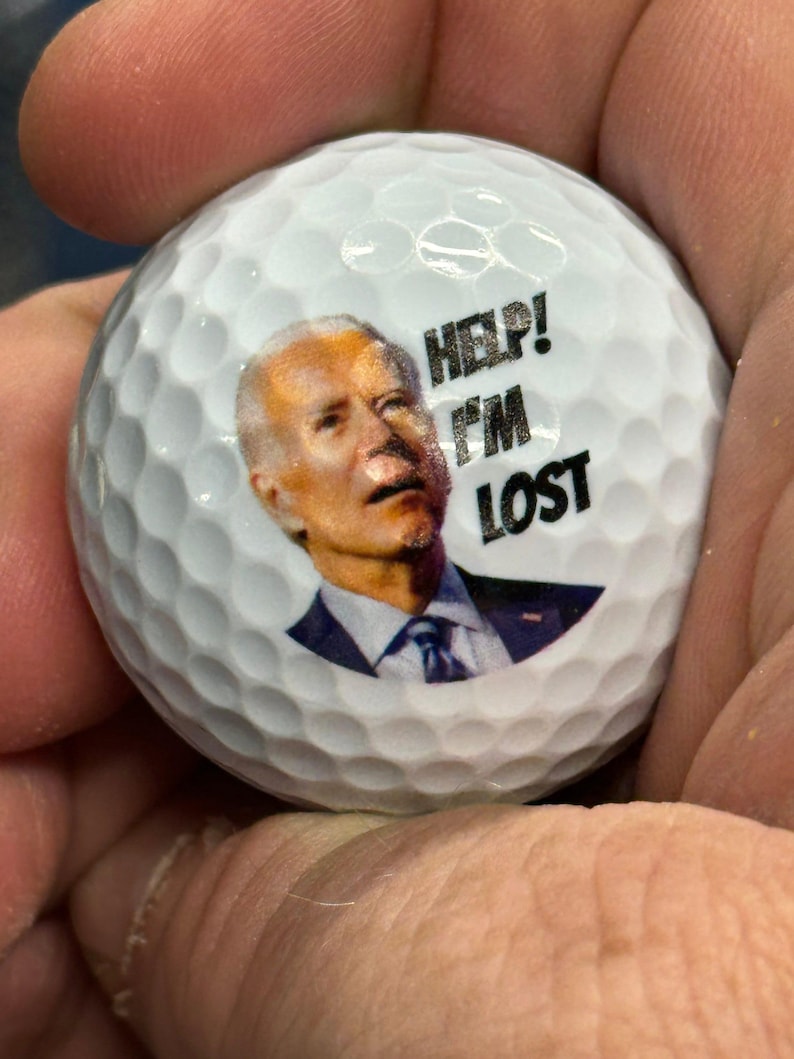 Puede incluir: Una bola de golf blanca con una imagen impresa de una persona con el texto "HELP! I'M LOST" en ella.
