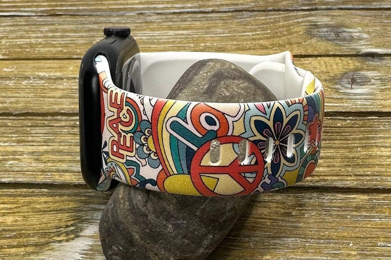 May include: A white Apple Watch band with a colorful psychedelic print featuring peace signs, flowers, and the word "Peace".