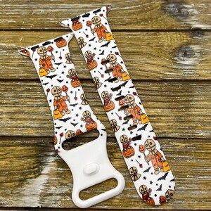 May include: White watch band featuring a Halloween-themed pattern. The design includes orange and brown figures, pumpkins, bats, and stars. The band has a white clasp and is set against a wooden background.