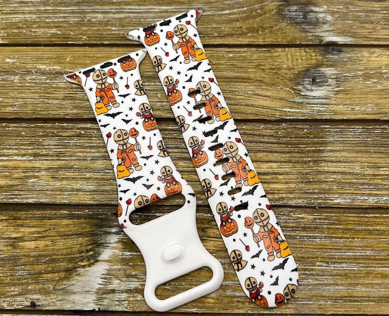 May include: White watch band featuring a Halloween-themed pattern. The design includes orange and brown figures, pumpkins, bats, and stars. The band has a white clasp and is set against a wooden background.