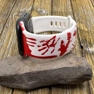 Puede incluir: Un reloj inteligente negro con una correa blanca con dise&ntilde;os de huellas de manos rojas. El reloj est&aacute; sobre una piedra gris sobre un fondo de madera. La correa tiene peque&ntilde;os agujeros para ajustar.