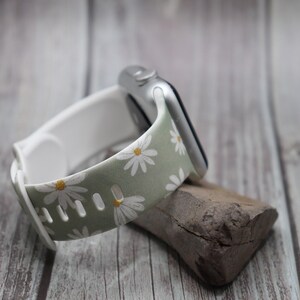 Puede incluir: Una correa de reloj con estampado floral verde y blanco con margaritas blancas. La correa est&aacute; hecha de silicona y es compatible con Apple Watch.
