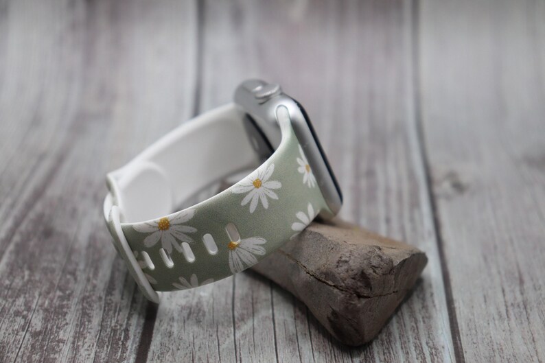 Puede incluir: Una correa de reloj con estampado floral verde y blanco con margaritas blancas. La correa est&aacute; hecha de silicona y es compatible con Apple Watch.