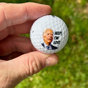 Puede incluir: Una bola de golf blanca con una imagen de un hombre con un traje azul y corbata. El texto "Help! I'm Lost" est&aacute; impreso en la bola.