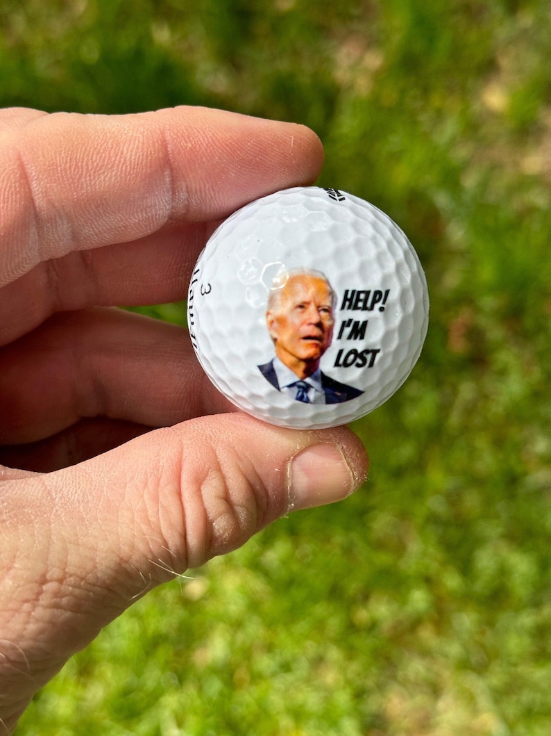 Puede incluir: Una bola de golf blanca con una imagen de un hombre con un traje azul y corbata. El texto "Help! I'm Lost" est&aacute; impreso en la bola.