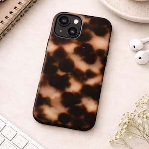 May include: A smartphone case with a tortoiseshell pattern in shades of brown and tan. The case is on a light-colored surface, with a keyboard, pen, notebook, and a cup of coffee in the background.