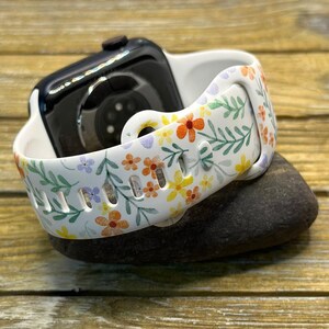 Op de afbeelding: Een wit horlogebandje met een bloemenpatroon met kleine gele, oranje en paarse bloemen en groene bladeren. Het bandje is gemaakt van siliconen en is compatibel met de Apple Watch.