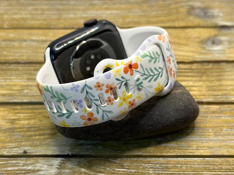 Op de afbeelding: Een wit horlogebandje met een bloemenpatroon met kleine gele, oranje en paarse bloemen en groene bladeren. Het bandje is gemaakt van siliconen en is compatibel met de Apple Watch.