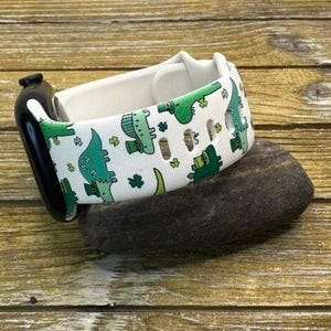 Puede incluir: Banda de reloj de silicona blanca con un estampado verde y blanco de dinosaurios duendes. La banda está sobre una superficie de madera marrón.