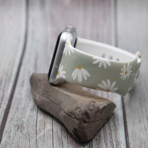 May include: A green and white floral watch band with a white daisy pattern. The band is made of fabric and has a silver metal buckle.