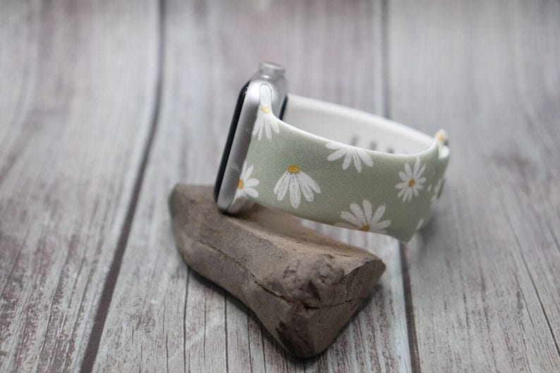 May include: A green and white floral watch band with a white daisy pattern. The band is made of fabric and has a silver metal buckle.