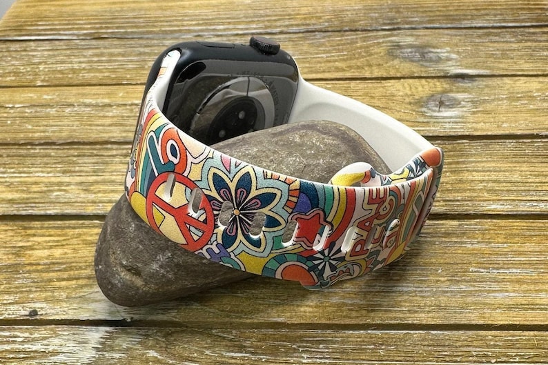 May include: White Apple Watch band with a colorful psychedelic pattern featuring peace signs, flowers, and rainbows. The band is made of silicone and has a smooth finish.