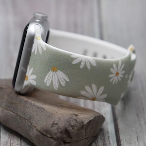 May include: A green and white watch band with a daisy print. The band is made of silicone and has a white lining.