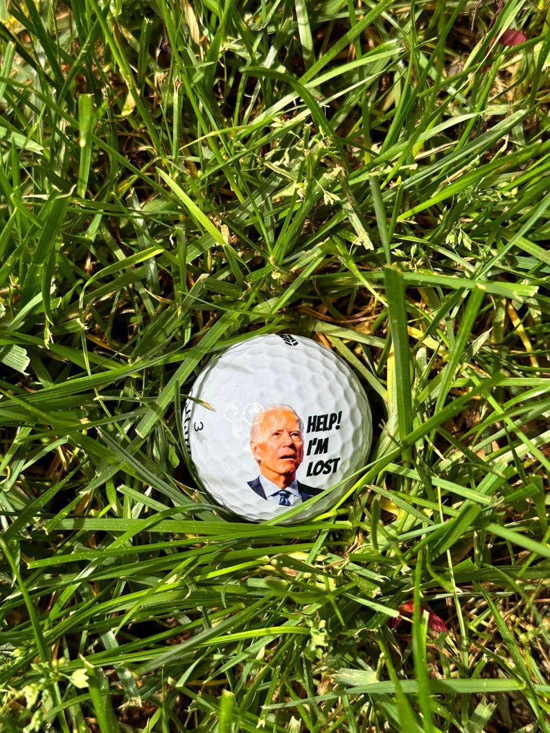 Puede incluir: Una bola de golf blanca con una imagen de un hombre en ella. El texto en la bola dice "Help! I'm Lost". La bola est&aacute; sentada en la hierba verde.