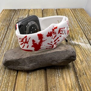 Puede incluir: Una correa de reloj de cer&aacute;mica blanca con dise&ntilde;os de huellas de manos y salpicaduras rojas, que sostiene un reloj inteligente negro. La correa descansa sobre una piedra gris, sobre un fondo de madera.