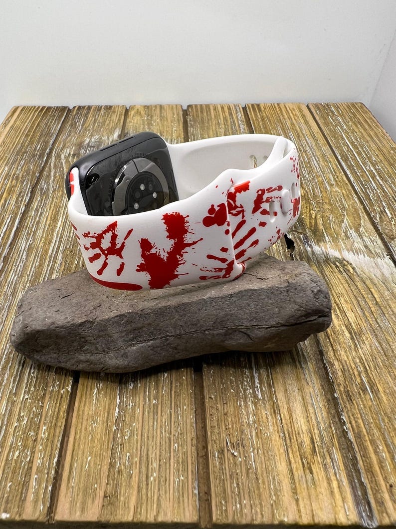 Puede incluir: Una correa de reloj de cer&aacute;mica blanca con dise&ntilde;os de huellas de manos y salpicaduras rojas, que sostiene un reloj inteligente negro. La correa descansa sobre una piedra gris, sobre un fondo de madera.