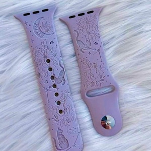 Op de afbeelding: Twee lavendelkleurige siliconen horlogebandjes met een gegraveerd ontwerp van katten, bloemen en sterren. De bandjes zijn ontworpen voor Apple Watches.