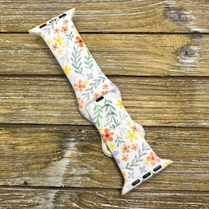 Op de afbeelding: Witte Apple Watch band met een bloemenpatroon met oranje, gele en paarse bloemen op een witte achtergrond.