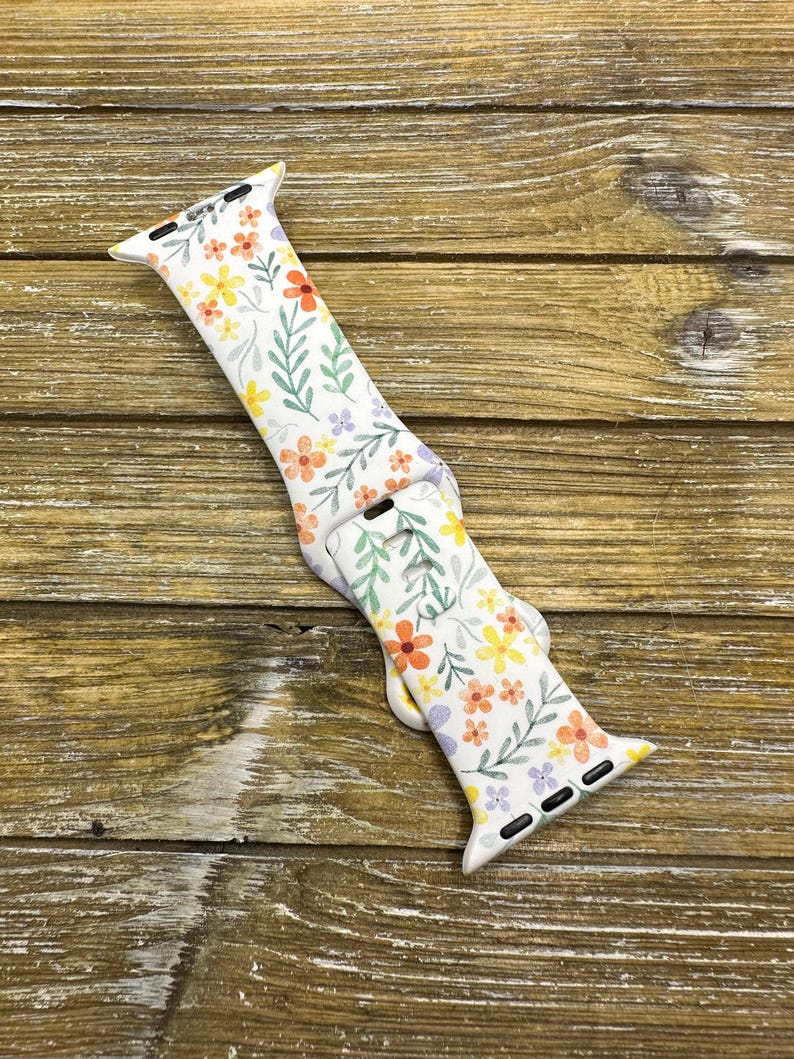 Op de afbeelding: Witte Apple Watch band met een bloemenpatroon met oranje, gele en paarse bloemen op een witte achtergrond.