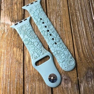 May include: A pair of light blue silicone Apple Watch bands with a sunflower design. The bands are shown on a wooden surface.