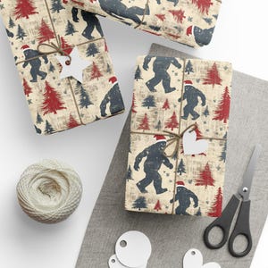 May include: Several wrapped gifts with Bigfoot and Christmas tree patterns in red and navy blue on a beige background. The gifts are tied with twine and adorned with white tags. A ball of twine and scissors are also present.