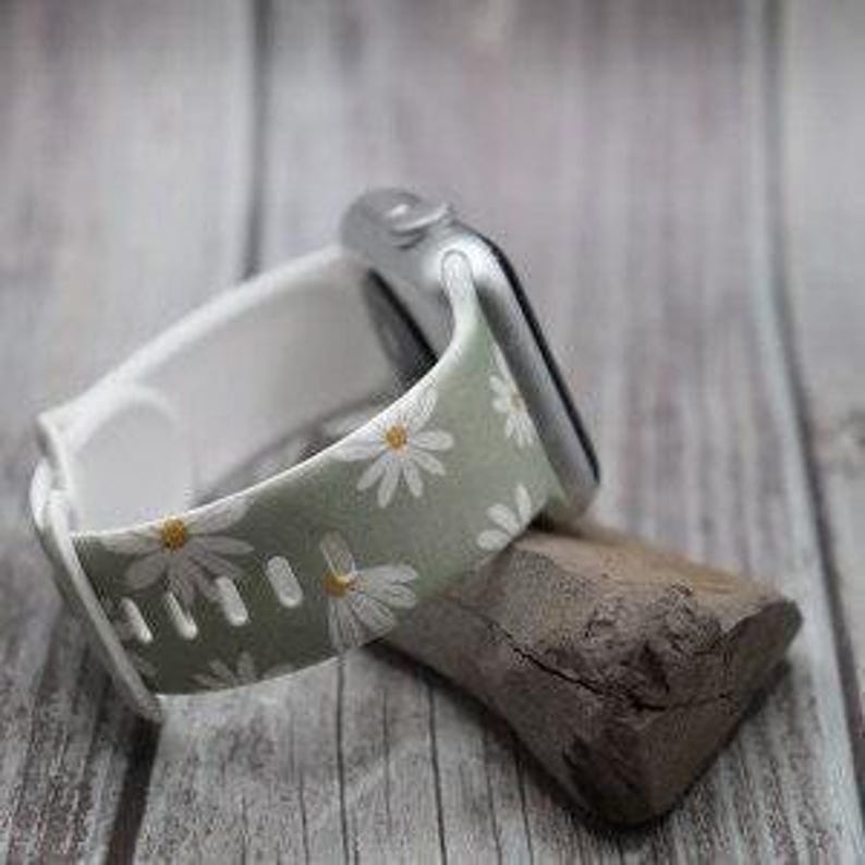 May include: A green and white Apple Watch band with a daisy print. The band is made of silicone and has a white buckle.