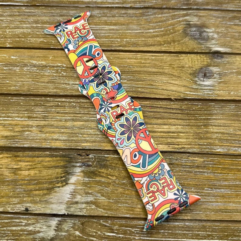 May include: A colorful Apple Watch band with a peace sign and "Peace" written in a retro font. The band is made of silicone and has a bright, psychedelic design.