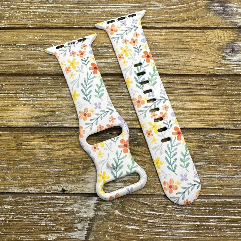 Op de afbeelding: Twee witte siliconen horlogebandjes met een bloemenpatroon. Het patroon bevat kleine gele, oranje en paarse bloemen met groene bladeren op een witte achtergrond.