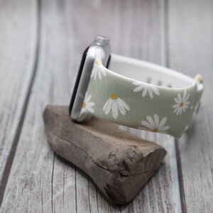 Puede incluir: Una banda de reloj Apple verde claro con estampado de margaritas blancas. La banda est&aacute; sobre una superficie de madera.