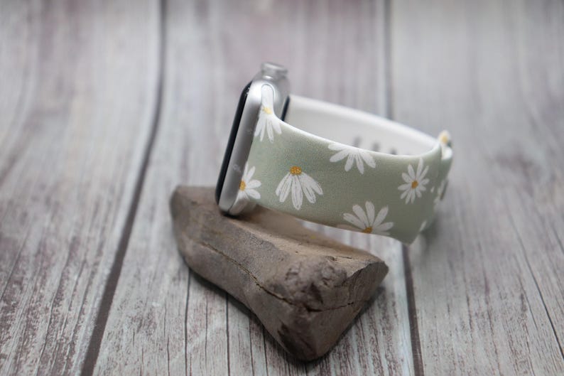 Puede incluir: Una banda de reloj Apple verde claro con estampado de margaritas blancas. La banda est&aacute; sobre una superficie de madera.