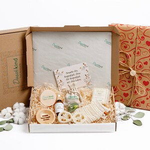 Può includere: Una scatola regalo piena di prodotti per il bagno e il corpo. La scatola è avvolta in carta marrone con cuori rossi. I prodotti includono un sapone, una bottiglia di olio per il corpo, una spugna vegetale e un panno. La scatola contiene anche un piccolo biglietto con il testo "Spezialle für Augenblicke, die für Augenblicke, die für das Leben sind."