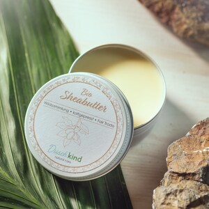 Può includere: Una latta bianca con un'etichetta marrone su cui è scritto "Bio Shea Butter" e "Wildsammlung - kaltgepresst - Fair Trade". L'etichetta presenta anche un disegno di un albero di karitè e il testo "Duschkind natural care".