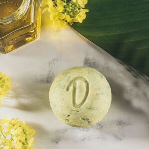 Può includere: Una saponetta di shampoo rotonda, di colore verde chiaro e bianco, con la lettera "D" in rilievo sulla parte superiore. La saponetta è appoggiata su una superficie bianca.